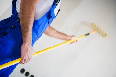 Close up, a builder in overalls paints a wall in white with a roller