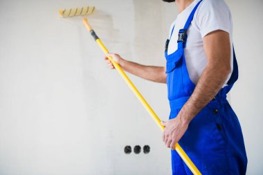 Close up, a painter in overalls paints a wall in white with a roller