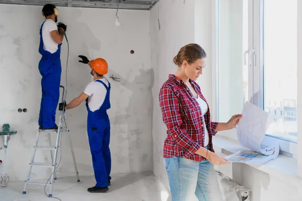The customer examines the layout of the apartment, two men are drilling the wall on a stepladder