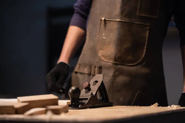 Un joven carpintero en un delantal trabaja en una carpintería, fabricando productos de madera. — Foto de Stock
