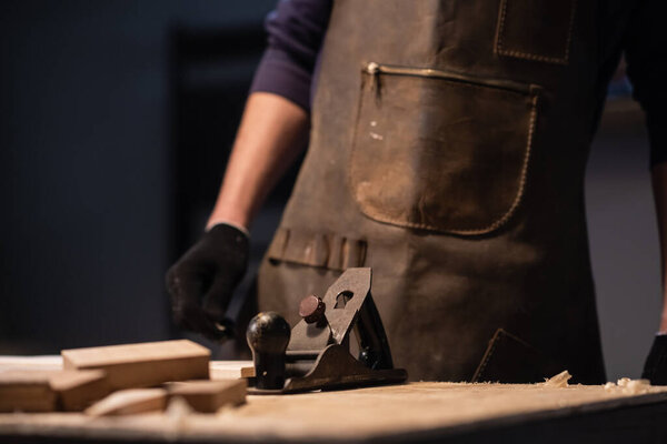 Молодой плотник в фартуке работает в столярной мастерской, изготавливая изделия из дерева.
