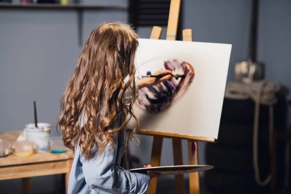 A young cute artist girl is learning to draw in an art workshop. Draws on an easel with oil paints and a brush. — Stock Photo, Image