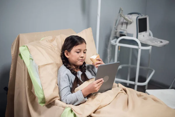 Niña positiva usando una aplicación en su dispositivo digital, mientras permanece en una cama de hospital — Foto de Stock