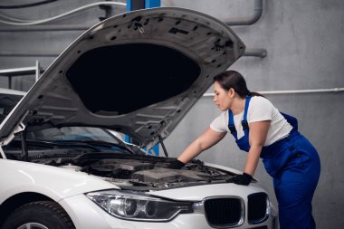 Kadın tamirci beyaz bir arabanın motorunu inceliyor.