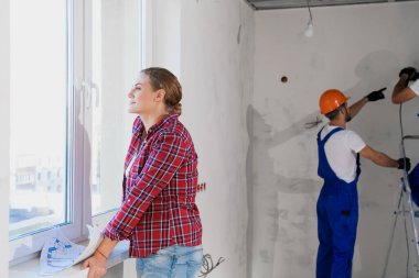 Bir kadın pencerenin yanında duruyor ve belgeleri inceliyor, arka plandaki inşaatçılar çalışıyor.