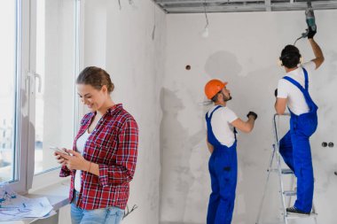 İnşaatçılar matkap ve merdiven kullanır, kadın müşteri telefonda yazar.