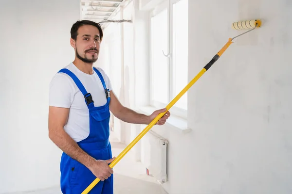 Sidovy av en ung manlig byggare måla en korridor vägg i vitt med en rulle — Stockfoto