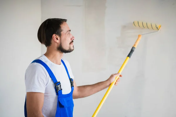 Man in blauwe overall schildert de muur met een roller, uitzicht vanaf opzij — Stockfoto