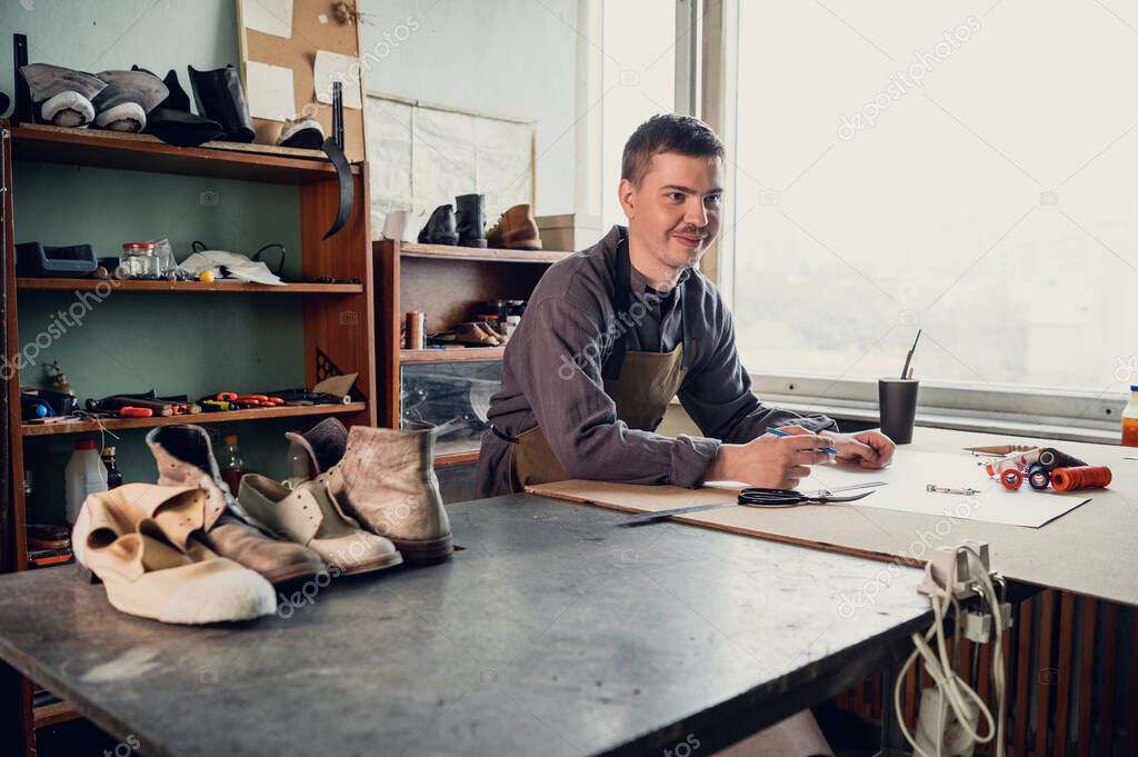 A young shoemaker makes a drawing for a pattern for leather shoes on a table in his workshop
