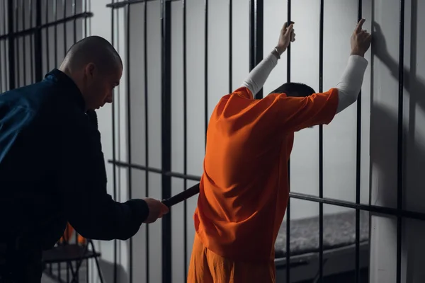 An armed guard searches a newly arrived criminal in a prison corridor against a backdrop of bars. — Stock Photo, Image