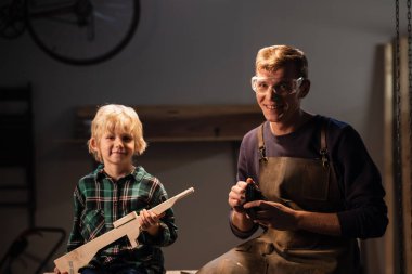 Genç bir marangoz baba ve yakışıklı sarışın oğlu ağaçtan bir silah yaptılar ve bunu gösterdiler, mutlular, marangozların atölyesinde masanın üstünde oturuyorlar..