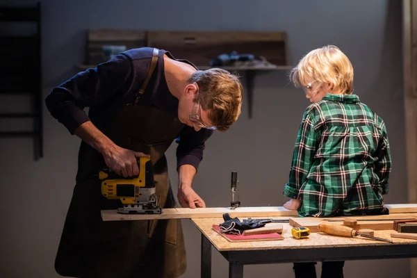 Un carpintero experimentado muestra el trabajo de varias herramientas a su hijo en el taller. — Foto de Stock