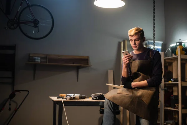 Un joven carpintero escribe un mensaje en su teléfono inteligente en un taller de carpinteros mientras está sentado en su escritorio a la hora del almuerzo. — Foto de Stock