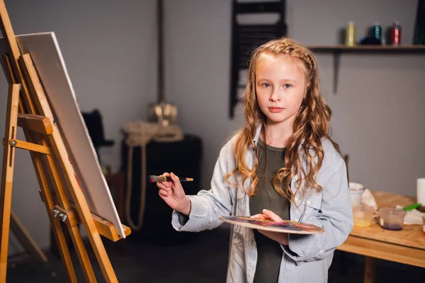 Una pequeña pintora posa y sonríe a la cámara cerca de su pintura en el estudio de artistas. — Foto de Stock