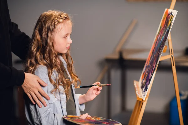 El padre apoya a la hija en aprender a dibujar. La maestra ayuda a la joven artista y sostiene sus manos sobre sus hombros. — Foto de Stock