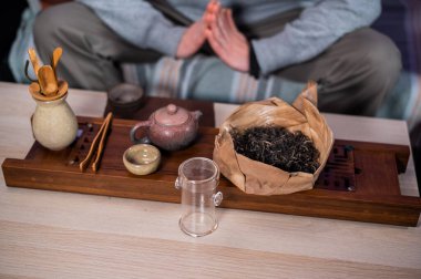 Shot without a face. A man meditates in front of a table with a full set of items for the tea ceremony, bamboo tray and tea in a clay teapot. Blurry background.