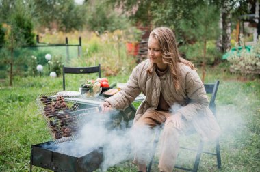 Karantinada sıkılan genç bir kadın ızgarada ızgara yapıyor. Yanında sebze, abur cubur ve bira olan bir masa var..