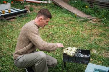 A young man in the backyard of the garden roasts sliced zucchini on the grill in the grill, next to a table with other vegetables