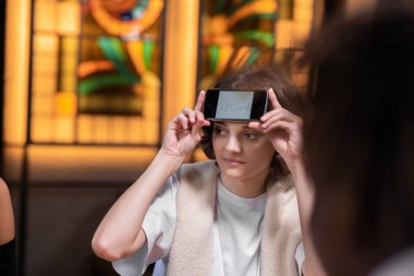 Alnında akıllı telefonu olan güzel bir kız arkadaşlarıyla masada oturuyor ve sergisine yazılmış bir kelimeyi tahmin etmeye çalışıyor, resmi kapat.