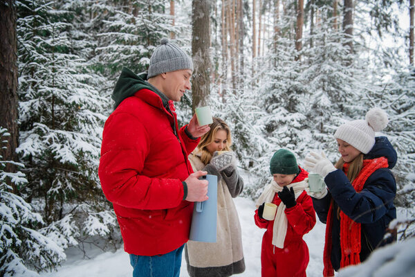 A young happy family with two children spends time outdoors in the winter forest. They drink hot tea from a thermos and mugs.