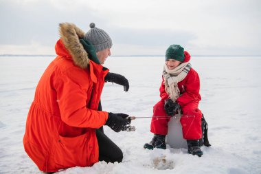 Kırmızı tulumlu ve eşarplı küçük bir çocuk babasıyla bir buz deliğinde balık tutuyor.