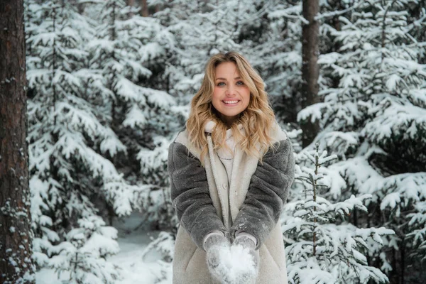 Счастливая блондинка с голубыми глазами выбрасывает снежинки из рук. Красивый портрет с боке в зимнем лесу. — стоковое фото