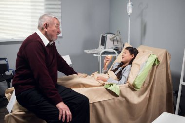 Son sınıf öğrencisi bir çocuğa hastanede bir hikaye anlatırken, çocuk yatakta kalıyor ve ilgiyle dinliyor.