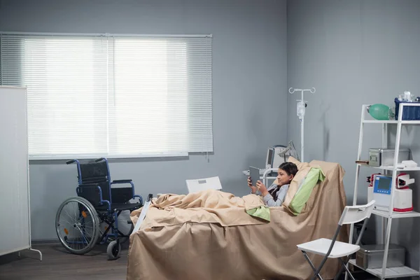 Uma menina descansando na cama em uma enfermaria do hospital, ela está jogando jogos em seu tablet digital — Fotografia de Stock