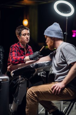 Yüzünde ve ellerinde piercingler ve dövmeler olan kısa saçlı bir kız, usta kızıl sakallı ve örgü şapkalı genç bir adam için bir dövme yapıyor..