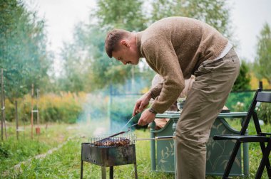 Koca, ailenin reisi, dışarıda bir akşam yemeği sırasında karısı için biftek ve barbekü hazırlıyor. Bir kır evinin bahçesindeki çimenlikte..