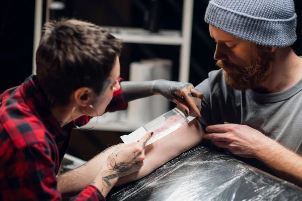 Chica artista del tatuaje se encarga de la mano de los jóvenes hombres después del tatuaje. Hace que la película transparente apósito con la aplicación de una crema hidratante. — Foto de Stock