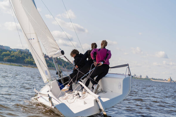 Быстрая, спортивная, одномачтовая яхта с тремя спортсменами на борту паруса с попутным ветром на красивой реке.