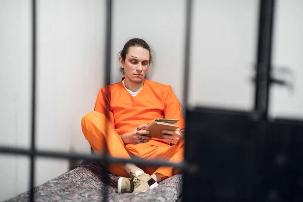 Ung brottsling i en isolerad cell läser en bok som sitter på en säng i en orange fängelseuniform. Foto genom staplarna. — Stockfoto