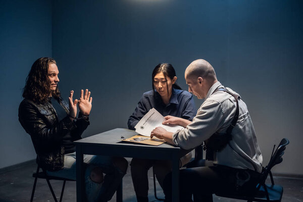 A tough police detective interrogates a crime suspect with a tattooed face in an interrogation room.