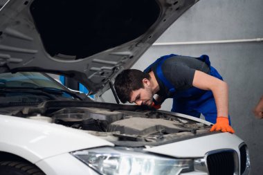 Bir tamirci arabanın yanında durur ve el hareketi gösterir.