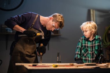 Genç bir erkek marangoz atölyesinde genç bir çocuğa marangozluk öğretiyor..