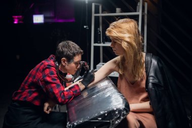 Modern deneyimli dövme ustası kızıl saçlı genç bir kızın koluna dövme yapıyor..