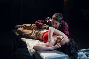 Kısa saçlı dövme ustası. Yüzünde ve ellerinde piercing ve dövmeler var. Kuaförde genç bir kadın için dövme yapıyor.