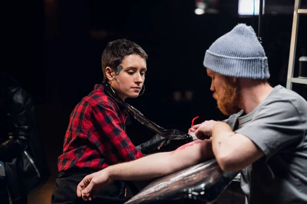 All'interno di un salone di tatuaggi, un giovane con la barba viene tatuato sul braccio usando il metodo stick e poke. Giovane artista ragazza in una camicia a scacchi. — Foto Stock