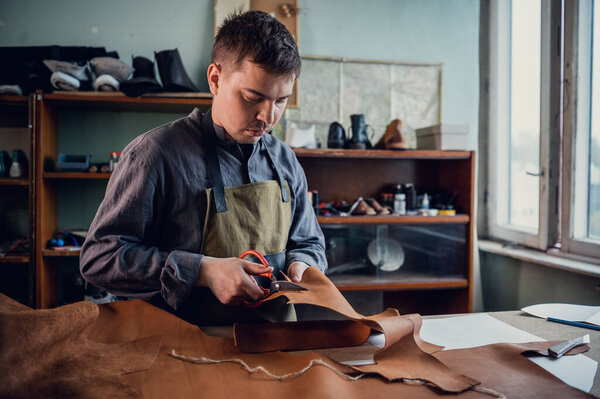 A young tanner in his workshop at the table cuts out leather elements of shoes