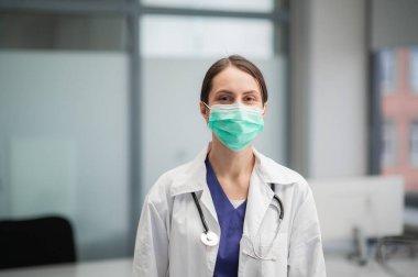 Şirin bayan doktor fotoğraf için poz veriyor, maskeli kameraya bakıyor.