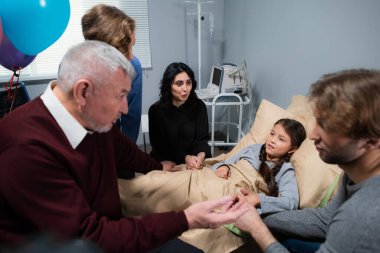Hastane koğuşundaki küçük bir kızın etrafında oturan ve konuşan bir aile.