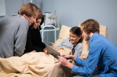 Küçük mutlu bir kız hastanede yatarken tablet kullanmayı öğreniyor. Ağabeyi de ona yardım ediyor.