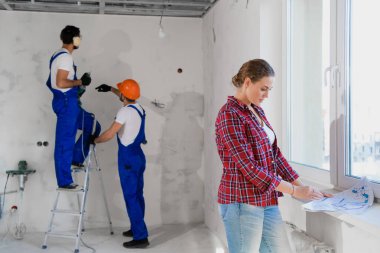 Müşteri, arka planda bir apartman dairesi planlamasını inceler. Elinde matkapla merdivenin üstünde durur.