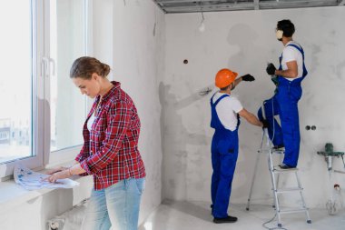 Bir kadın bir apartman dairesinin planlarını inceliyor, arka plandaki inşaatçılar elinde matkapla bir merdivende duruyorlar.