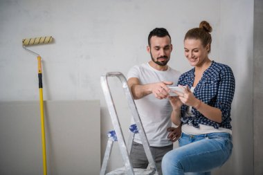 Bir adam ve kız arkadaşı bir apartman dairesinde yenilenen akıllı telefondan bir fotoğrafa bakıyorlar.