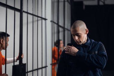 Tam teçhizatlı bir gardiyan telsizdeki ortağıyla iletişim kuruyor. Arka planda, hücrede bir mahkum.