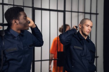 Siyah ve beyaz gardiyanlar, arka planda mahkumlarla birlikte batonlarıyla dururlar..