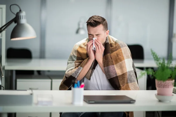 Young man gets sick at work and blows his nose with a napkin at the office desk