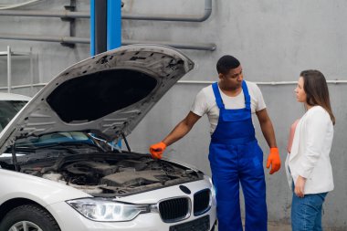 Tulumlu tamirci ve müşteri arabanın motorunu inceliyor.